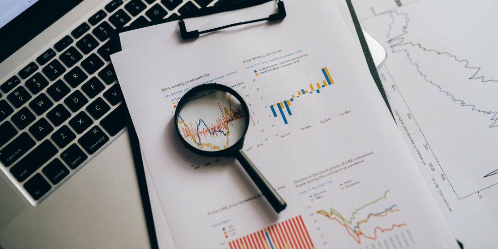 A laptop and papers showing business finances, along with a magnifying glass. This signifies close analysis of business finance data.